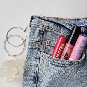Jeans pocket with lip balms, hoop earrings, and a floral accessory on white.